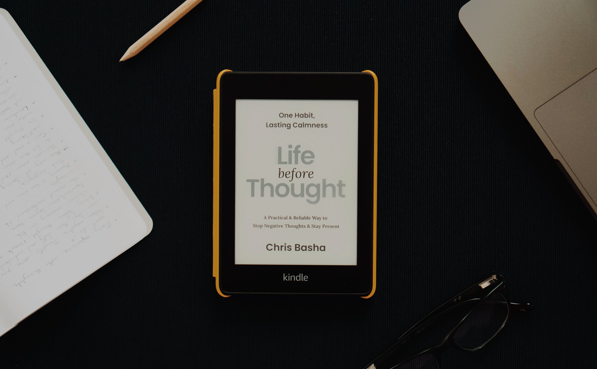 Life Before Thought open on the title page of a Kindle, on a dark desk with a notebook, pencil, laptop, and glasses
